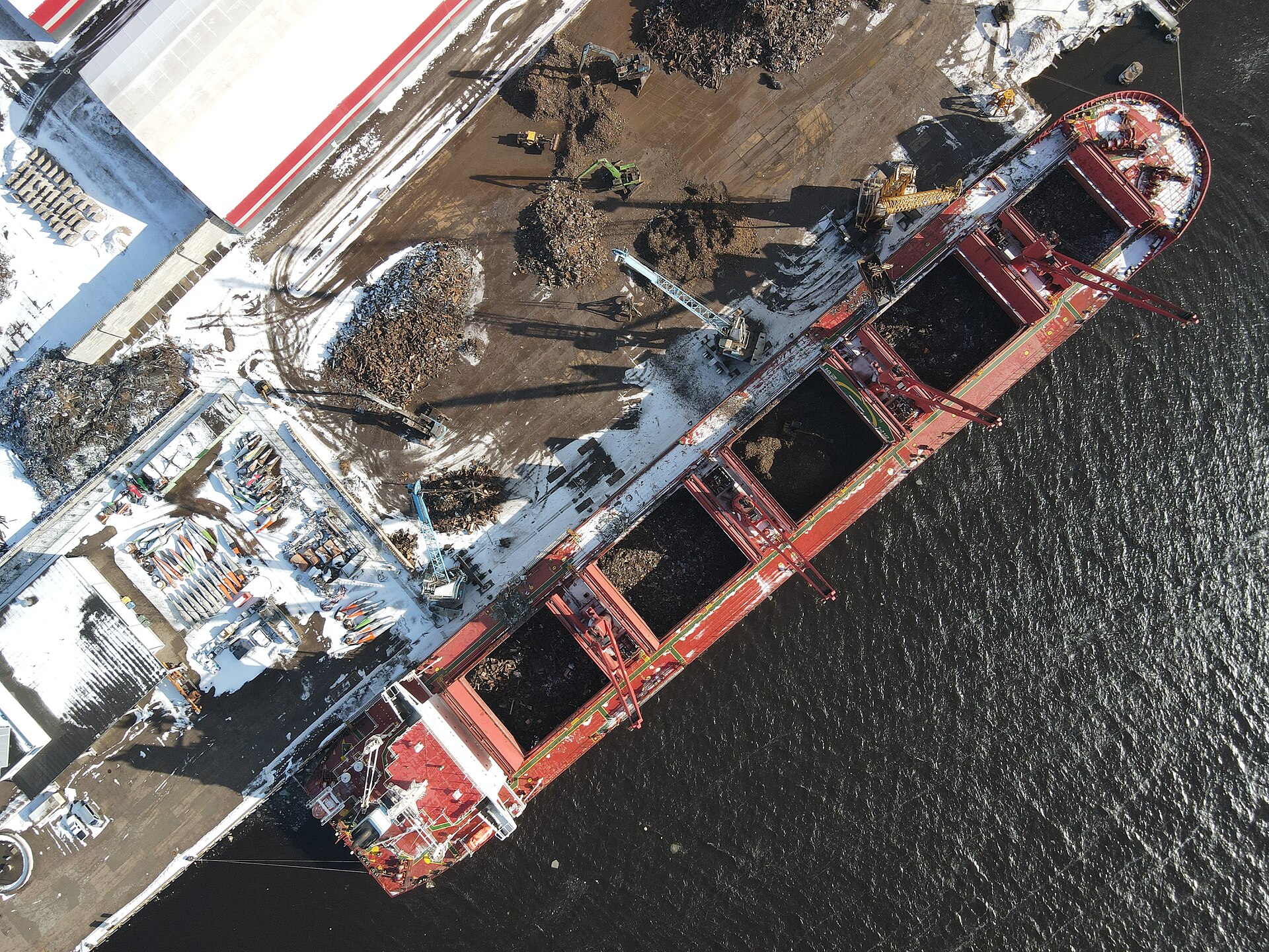 Overhead documentary view of a geared bulker handling metal scrap at a snow-covered quay, with open holds and shore grabs.